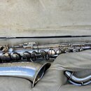 Stříbrný saxofon v otevřeném kufru s vnitřním polstřením. Na čáře nástroje je vidět nápis „S. MAUQUOT 22“ a pod ním texty „PARIS“, „Série No. 3549,5“…