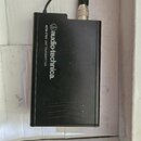 Černý obdélníkový vysílač s připojeným kabelem a kovovým konektorem. Na přední straně je bílý nápis „audio-technica.“ s logem a vedle něj text „ATW-T…