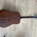 Akustická kytara v tmavě hnědé barvě s viditnou strukturou dřeva. Nástroj leží na světlé dřevěné podlaze. Na hlavě kytary jsou vidět stříbrné ladičky…