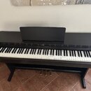 Černý digitální piano s klaviaturou a stojanem. Uprostřed nad klávesami je bílý nápis YAMAHA. Nástroj je vybaven třemi kovovými pedály a hudebním pul…