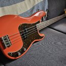 Oranžová elektrická basová kytara s tmavě hnědým pickguardem s texturou želvy. Na hlavě krku je viditelné logo "Fender". Nástroj má čtyři struny, dva…