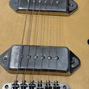 Detail korpusu elektrické kytary z přírodního světlého dřeva. Jsou vidět dva chromované humbuckery a most s šesti sedly. Povrch kovových částí vykazu…