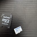 Na fotografii je černý adaptér a malá paměťová karta microSD. Adaptér nese nápisy Lock, aerbes®, microSD a Adapter s označením CE. Karta je bílá s mo…