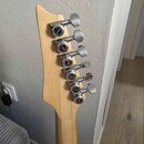 Detail hlavy světlé dřevěné kytary s šesti chromovanými ladicími mechanismy. Na každém mechanismu je viditelný nápis "M BUTTON". Povrch dřeva i kovov…