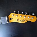 Fotografie zachycuje hlavu elektrické kytary z žlutého dřeva s šesti kovovými ladičkami. Na povrchu je černý nápis Fender® TELECASTER a pod ním menší…