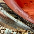 Detailní záběr na mosazný povrch saxofonu s lakem. Na těle nástroje je vyryta dekorativní ornamentika a nečitelný nápis. Povrch vykazuje drobné škráb…