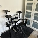 Elektronická bicí souprava s černým rámem a padami. Na sedáku je bílý nápis Pearl. Souprava obsahuje čtyři pady, dva činely a pedály, vedle ní stojí…