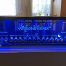 Kytarový zesilovač s černým šasi a modrým podsvícením, v jehož horní části jsou vidět elektronické lampy. Na předním panelu je bílý nápis Hughes & Ke…
