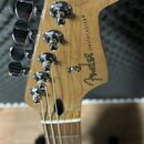 Detail hlavy elektrické kytary z přírodního dřeva s chromovanými ladičkami. Na dřevě je vidět nápis "Fender STRATOCASTER" a v horní části text "ORIGI…