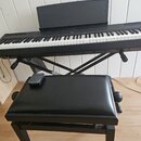 Černý digitální klavír na černém X-stojaně s přiloženým černým polstrovaným sedákem. Na klavíru je nápis YAMAHA a na dálkovém ovladači položeném na s…