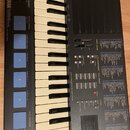 Černý elektronický klavír s bílými a černými klávesami. Na levé straně je nápis YAMAHA Perfume, pod ním text PGM-100 / DSS / AWM2 / FM VOICE BANK. Př…