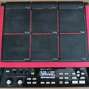 Černý sampler s devíti gumovými pady a červeným rámem. Na spodním ovládacím panelu je bílý nápis Roland SP-90 SX. Zařízení obsahuje displej s textem…