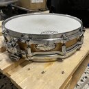 Na fotce je malý snare drum s dřevěným korpusem v přirozené hnědé barvě a chromovaným kováním. Na korpusu je viditelný oválný štítek s nápisy „Pearl“…