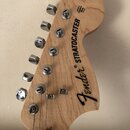 Fotografie zachycuje hlavu elektrické kytary z přírodního světlého dřeva. Na povrchu je černý nápis Fender® STRATOCASTER. Jsou vidět chromované ladič…