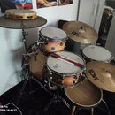 Bubnová sada s přirozeným dřevěným povrchem v oranžovém odstínu. Na tomu a snare bubnu je viditelné logo Pearl, na tomu bubnu je navíc nalepený kulat…