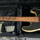 Elektrická basová kytara krémové barvy s černým pickguardem v černém plišovém kufru. Na hlavě hmatníku je viditelné logo Fender. Tělo nástroje vykazu…