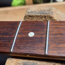 Detail hmatníku strunového nástroje z tmavého dřeva s viditnou strukturou slz. Na hmatníku jsou kovové pražce a dvě bílé kulaté polohové značky. Povr…