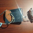 Fotografie zobrazuje elektrický snímač kytary s připojenou černou plastovou základnou a vodiči. Na bílém štítku u vodičů je částečně viditelný nápis…
