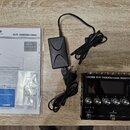 Na fotografii je černý kytarový procesor s nápisem BOSS GT-1000CORE GUITAR EFFECTS PROCESSOR. Vedle přístroje je kladen černý síťový adaptér a uživat…
