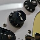 Detail elektrické kytary v žluté barvě s bílým pickguardem. Na snímku jsou vidět dva černé oválné knofíky s bílými nápisy VOLUME a TONE a číslováním…