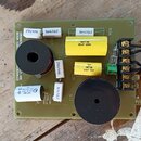 Fotografie znázorňuje elektronickou součástku, pravděpodobně crossover, na zelené desce plošných spojů. Na desce jsou umístěny dva černé cívky, bílé…