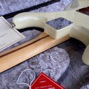 Detail zadní části kytary v krémové barvě s viditelným kovovým mechanismem u krku. Na kovovém krytu je vyraženo logo Fender. Vedle nástroje leží hněd…