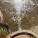 Detail stříbrného, kovového povrchu s jemnými rýhami a odrazem světla. Uprostřed je vyraženo kulaté logo s nápisem „HAND MADE IN SWITZERLAND“ v horní…