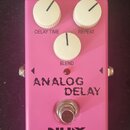 Růžový efekt pedál Analog Delay s třemi bílými oválnými knoflíky označenými jako DELAY TIME, REPEAT a BLEND. Na spodní části je nápis ANALOG DELAY, l…