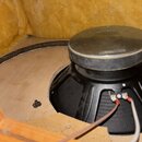 Fotografie zachycuje zadní část černého reproduktoru s kovovým magnetem, který je osazen v dřevěné konstrukci. K reproduktoru vedou dva elektrické vo…