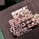 Černý síťový adaptér s bílým textem. Je na něm uveden nápis SHURE® AC ADAPTER, MODEL NO: PS31E, INPUT: 230V ~ 50Hz 0.06A a OUTPUT: 15V = 400mA. Dále…