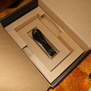 V otevřené hnědé kartonové krabici leží černý USB kabel. V pravém dolním rohu je vidět část černého elektronického zařízení s bílými nápisy VOLUME, S…