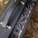 Detail ovládacího panelu černého kytarového komba. Panel obsahuje čtyři otočné potenciometry s popisky GAIN, TREBLE, BASS a VOLUME a přepínač s nápis…
