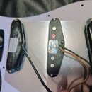 Fotografie zachycuje zadní stranu snímačů elektrické kytary s bílým tělem. Jsou zde tři černé snímače s viditelným zapájkováním a připojenými kabely.…