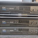 Tři černé rackové výkonové zesilovače značky QSC AUDIO. Horní jednotka nese nápis RMX 1850HD PROFESSIONAL POWER AMPLIFIER, prostřední RMX 1450 PROFES…
