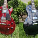 Dvě fotografie elektrických kytar typu Les Paul, jedna v červené a druhá v černé barvě. Obě mají dva humbuckery, tmavý hmatník a černý plechtrpickgua…