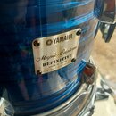 Detail modrého bubnu s kovovým štítkem připevněným čtyřmi šrouby. Na štítku je uveden nápis YAMAHA, Maple Custom DEFINITIVE a MADE IN JAPAN. Ve spodn…