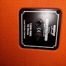 Detail zadní strany kytarového boxu s oranžovým potahem. Na černém plastovém panelu jsou dva vstupy a nápisy: „ORANGE VOICE OF THE WORLD“, „Serial No…