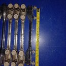 Fotografie zobrazuje pět kovových částí, pravděpodobně mechanismů pro ladění nebo držáků, položených vedle sebe na modrém povrchu. Čtyři z nich jsou…
