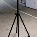 Černý kovový stativ s teleskopickým středovým sloupem a klikou pro regulaci výšky. Nástroj stojí na betonové podlaze v garáži. Na předmětu nejsou vid…