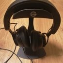Černé uzavřené sluchátka umístěná na černém stojanu s logem K&M. Na vršku náušnikového oblouku je nápis ADAM AUDIO. Sluchátka jsou připojena k černém…