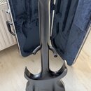 Zadní pohled na černou elektrickou kytaru s matným povrchem, umístěnou na černém stojanu před otevřeným kufrem. Na zadní straně hlavy jsou viditelné…