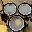Fotografie zobrazuje části elektronické bicí sady. Jsou vidět čtyři bílé padů s černými okraji, na kterých je nápis „Roland Reg. U.S. Pat. & Tm. Off.…