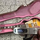 Fotografie zachycuje elektrickou kytaru v hnědém kufru s růžovým vnitřkem. V popředí je digitální váha značky eta Travello, která ukazuje hodnotu 4.0…