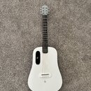 Bílá akustická kytara s matným povrchem a černým hmatníkem. Na hlavě je nápis "Lava Me". Tělo má netradiční tvar s oválným otvorem pro zvuk umístěným…