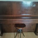 Tmavě hnědé dřevěné piano s uzavřenou klaviaturou a přívodní židličkou na třech nohách. Povrch nástroje vykazuje známky opotřebení v podobě odřeni a…