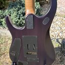 Zadní strana elektrické kytary v tmavě fialové barvě s dřevěným hnedým krkem. Na spojení krku a těla je kovový štítek s logem a nápisem "Music Man" a…