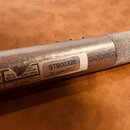 Detail stříbrného kovového trubkovitého předmětu s texturovaným povrchem v pravé části. Na těle je nalepený bílý štítek s nápisy STB00320 a 01 June 2…