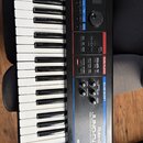 Černý digitální klavír s bílými a černými klávesami. Na předním panelu jsou nápisy Roland, JUNO-Di a MOBILE SYNTHESIZER WITH SONG PLAYER. Přístroj ob…
