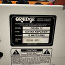 Fotografie detailu zadní strany kytarového komba s bílým povrchem. Na černém štítku je uvedeno logo ORANGE, nápis LONDON | ENGLAND VOICE OF THE WORLD…