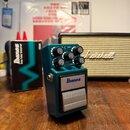 Modrý efekt pedál s nápisem Ibanez Bass Tube Screamer. Na horní části jsou čtyři ovládací knoflíky s popisky DRIVE, LEVEL, MID a TREBLE. Vedle pedálu…